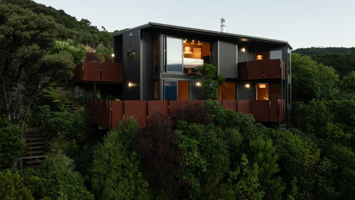 Golden Bay House nestled in steep bush at dusk, dark cladding and corten balconies lit from within.