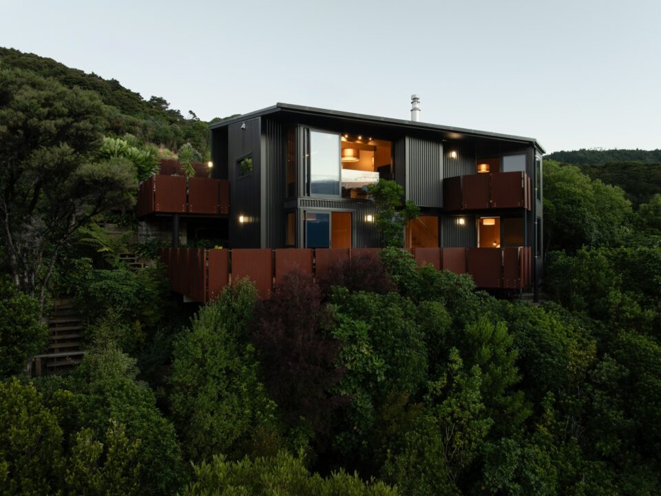 Golden Bay House nestled in steep bush at dusk, dark cladding and corten balconies lit from within.
