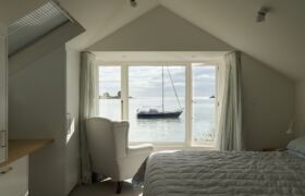 Bedroom with gable ceiling and armchair, framed by a wide window overlooking the estuary and a sailboat.