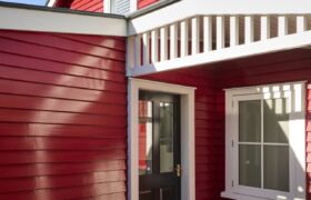 Red weatherboard entry with black front door, white trim and patterned tile threshold in afternoon sun.