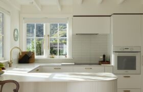 Bright white kitchen with brass tapware, tiled splashback and exposed rafters, lit by afternoon sun.
