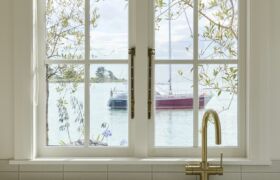 Brass mixer tap and white sink beneath a timber-framed sash window looking out to the estuary and moored boats.