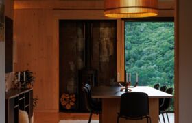 Dining space with timber ceiling, wood burner and pendant light, framed by bush-valley glazing.