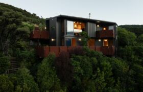 Golden Bay House nestled in steep bush at dusk, dark cladding and corten balconies lit from within.